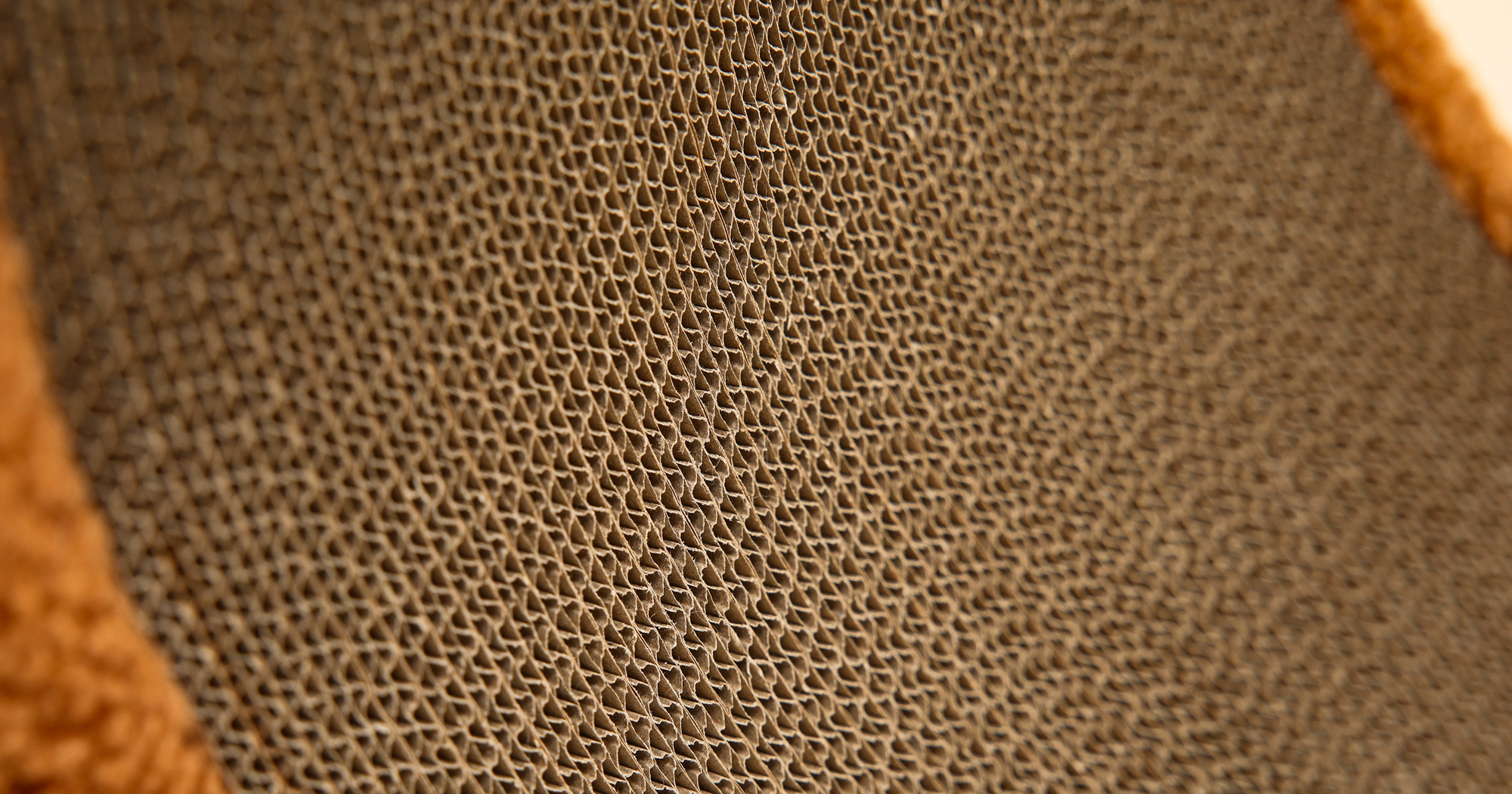 A macro photograph showcasing the dense, vertical corrugated cardboard pattern of our cat scratcher, with a soft, brown plush edge.