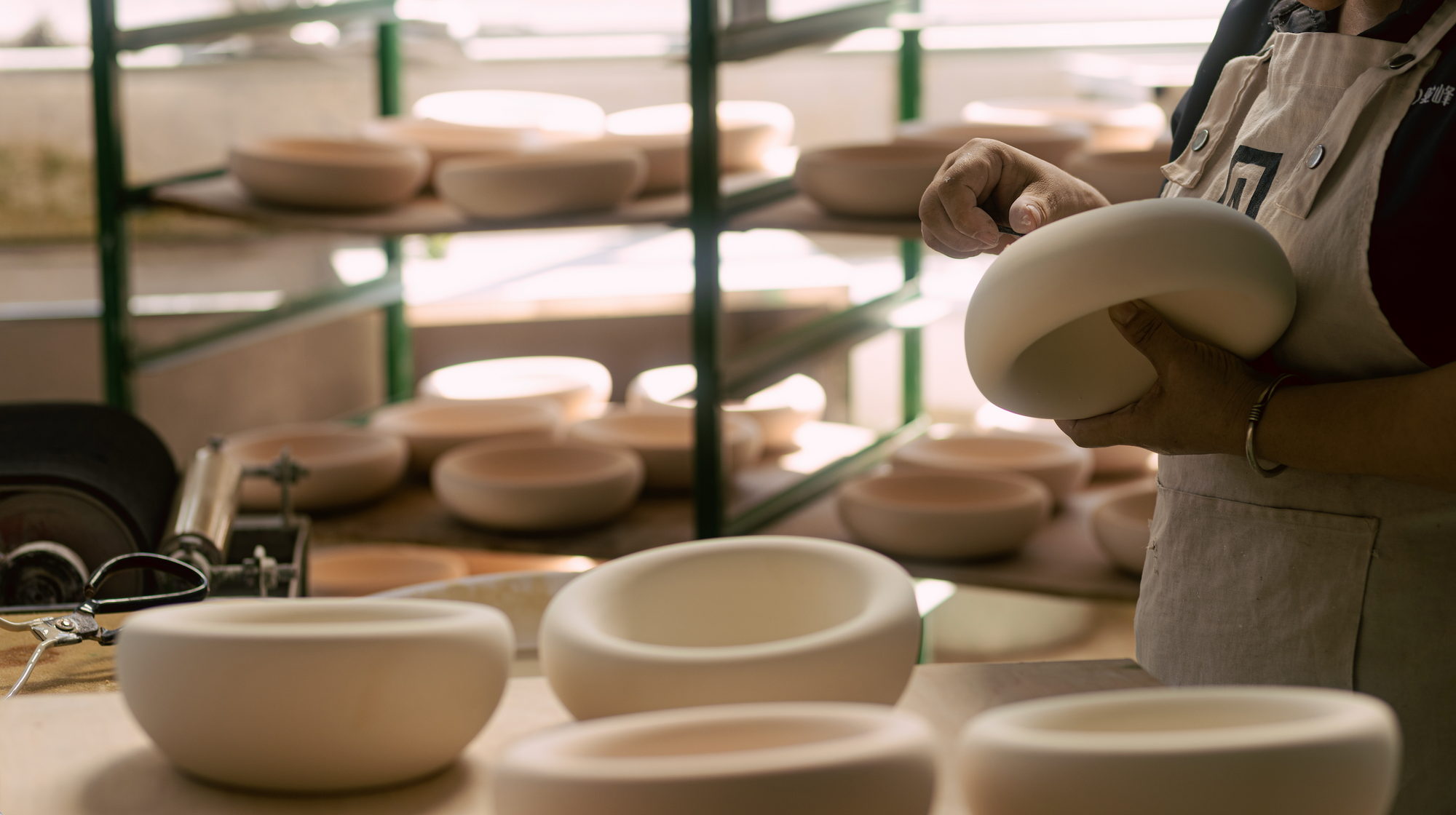 Artisan in a workshop hand-finishing a white ceramic pet bowl, with rows of unglazed Pet Jojo dog and cat bowls drying on shelves